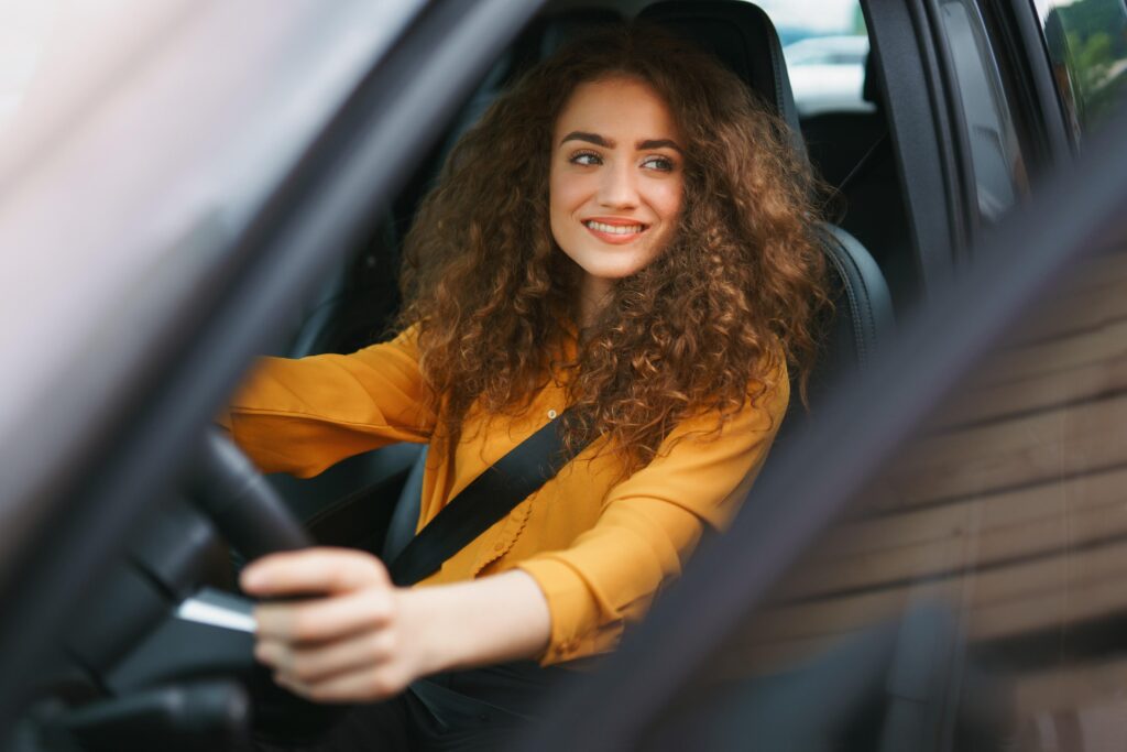 Smiling woman with dark, curly hair wearing a yellow blouse behind the wheel of a car