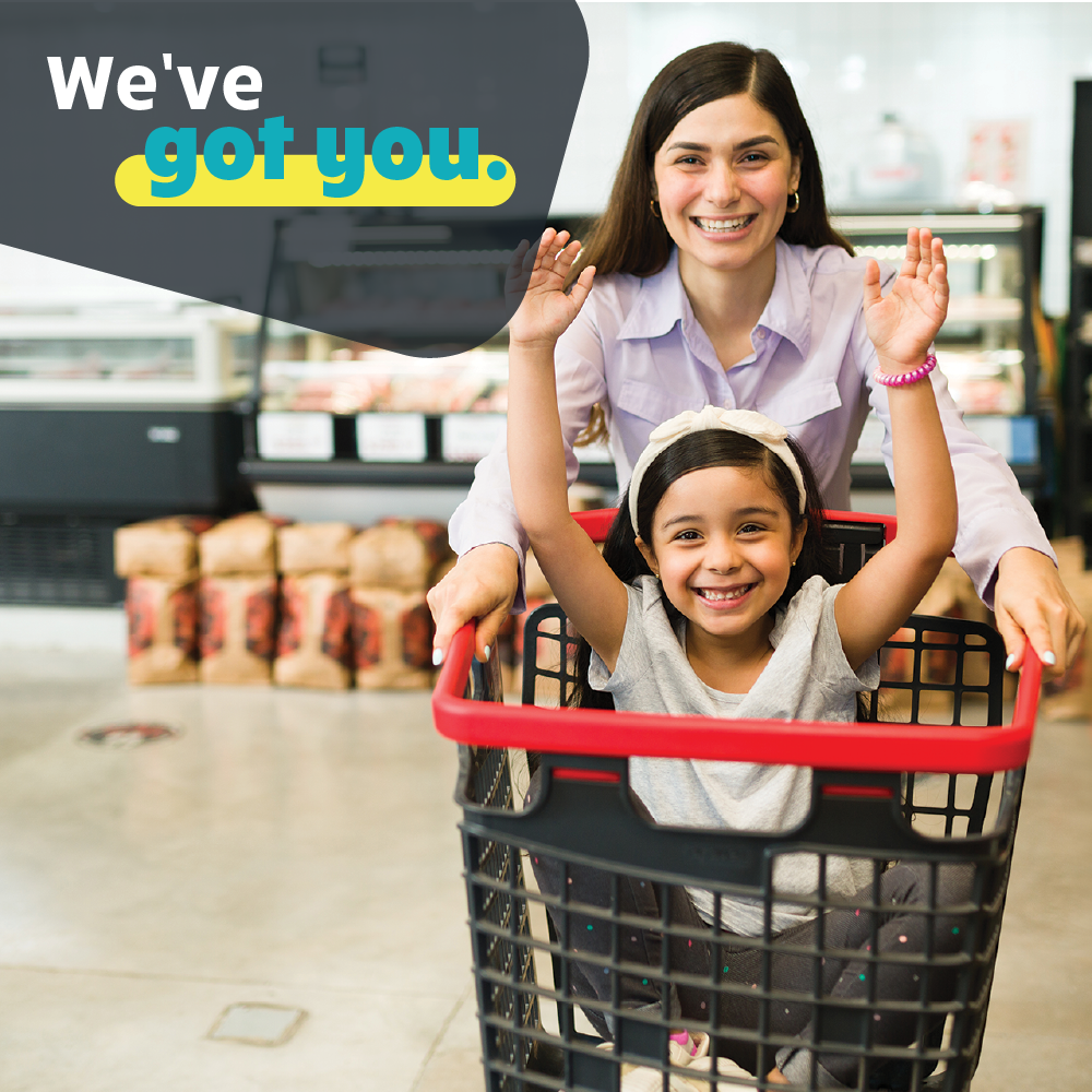 Photo of a mother pushing her young daughter in a grocery cart. The daughter's hands are in the air and she is smiling.