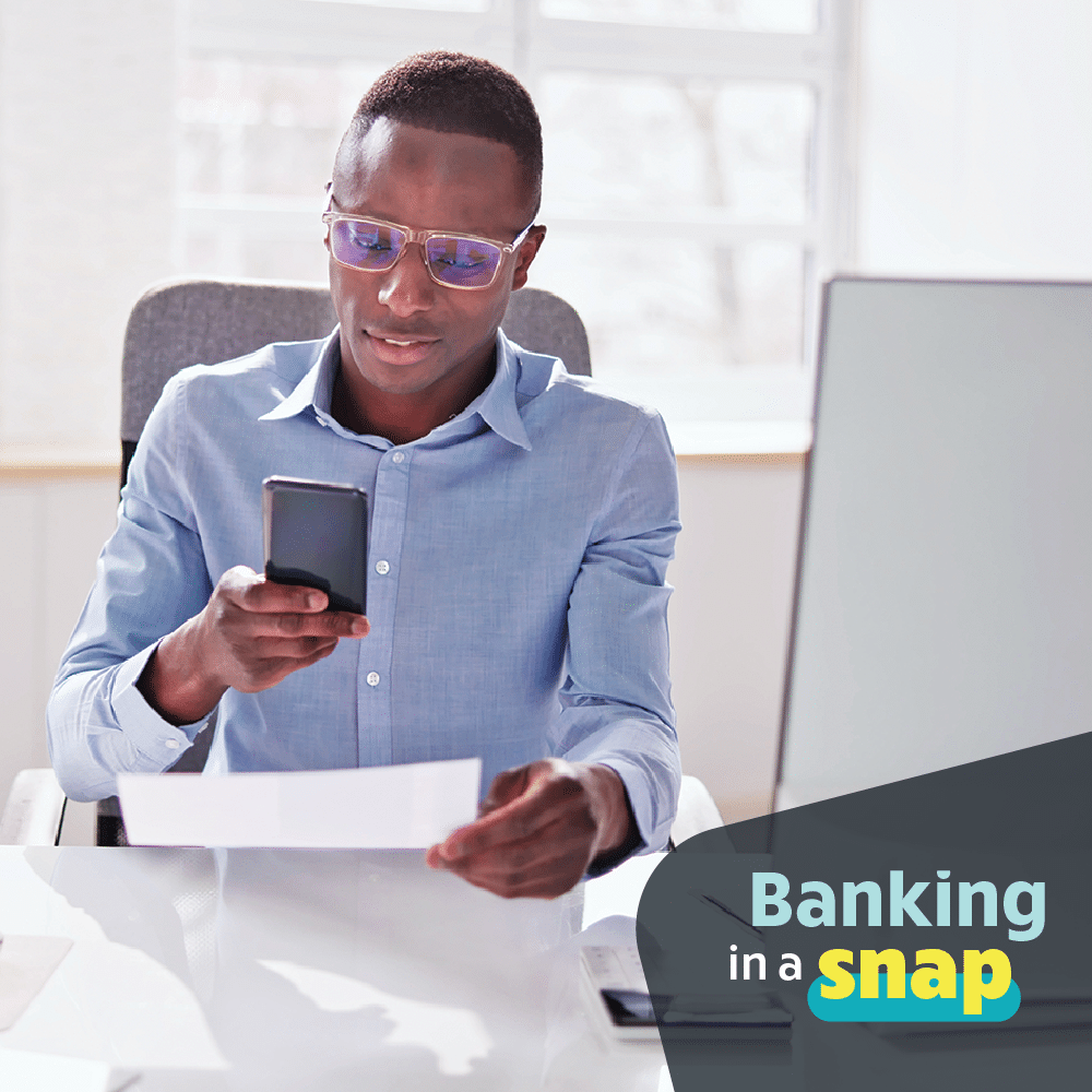 Photo of man in his 30s depositing a check with his phone while sitting at a desk in a pale blue shirt and eye glasses