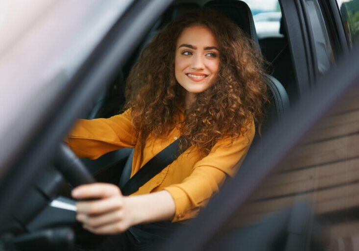 Smiling woman with dark, curly hair wearing a yellow blouse behind the wheel of a car