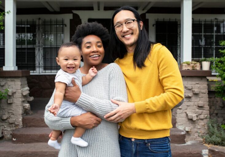 Happy couple with their infant standing in front of their home